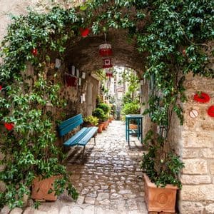 Una vista attraverso un arco in pietra coperto di viti in un vicolo stretto di ciottoli con una panchina blu e piante in vaso.