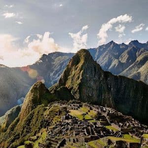 Antiguas ruinas de piedra se asientan en una ladera montañosa verde y aterrazada, dominadas por picos escarpados bajo un cielo nublado y soleado.
