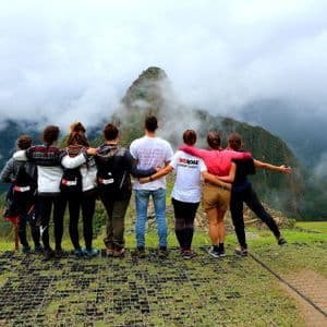 Un grupo de WeRoad, de pie y abrazados, en un mirador de hierba, contempla un pico de montaña cubierto de nubes.