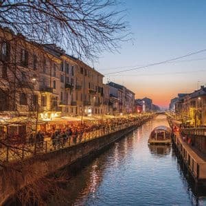 Un largo canal de ciudad al atardecer, con restaurantes iluminados y gente paseando a lo largo de la orilla del agua bajo un cielo colorido.