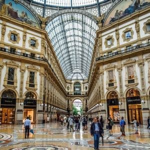 La gente pasea por una galería comercial ornamentada con un techo abovedado de cristal, intrincados suelos de mosaico y escaparates de lujo.