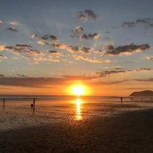 Un vibrante sole arancione tramonta sull'oceano, creando sagome di persone sulla sabbia bagnata e un riflesso sull'acqua.