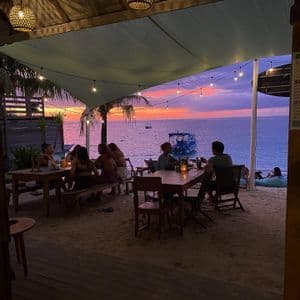 Un gruppo WeRoad cena in un ristorante sulla spiaggia sabbiosa, sotto le lucine, ammirando un vivace tramonto sull'oceano.