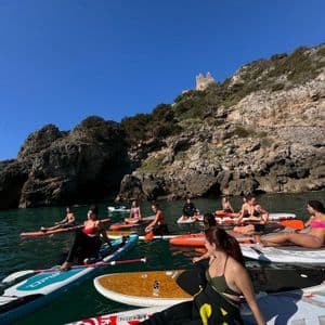 Un viaggio di gruppo WeRoad si rilassa su stand-up paddleboard nel mare, accanto a una scogliera rocciosa con rovine in cima.