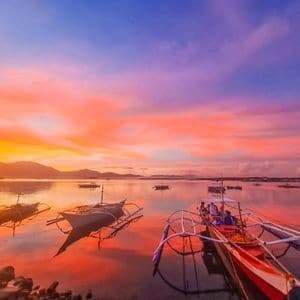 Barche a bilanciere galleggiano su acque calme sotto un tramonto vibrante arancione e viola, con montagne in lontananza.
