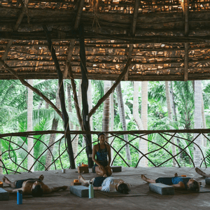 Un viaggio di gruppo WeRoad pratica yoga in un padiglione nella giungla a cielo aperto, con i partecipanti sdraiati sui tappetini e un istruttore inginocchiato.