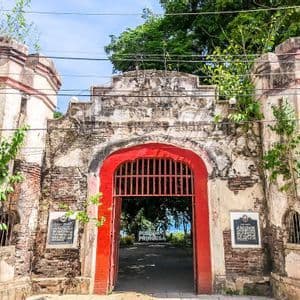 Un portone di fortezza in pietra logorata, con un ingresso ad arco rosso vivo e aperto, invaso da piante verdi.