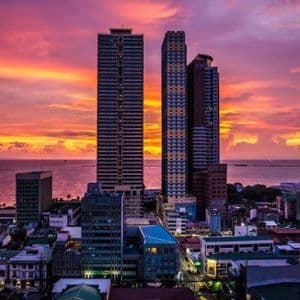 An aerial view of a coastal city's skyscrapers set against a vibrant orange and purple sunset over the ocean.