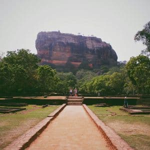 Un long chemin à travers les ruines d'un ancien jardin mène à une immense forteresse rocheuse, avec quelques personnes debout à sa base.