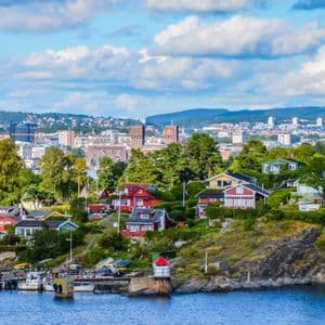 Des maisons colorées parsèment un littoral vert et rocheux devant une ligne d'horizon urbaine lointaine sous un ciel partiellement nuageux.