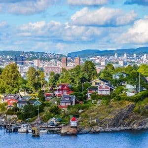Colorful houses on a rocky, tree-covered coastline with a city in the background under a blue sky with white clouds.