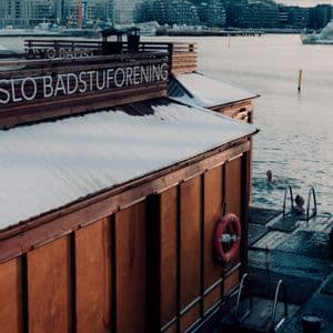 Zwei Menschen baden in kaltem Wasser neben einer schwimmenden Holzsauna, im Hintergrund eine schneebedeckte Stadtkulisse.