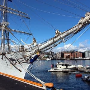 The bow of a large white tall ship with intricate rigging docked in a city harbor under a bright blue sky.