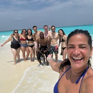 Une femme en bikini bleu prend un selfie souriant avec son voyage de groupe WeRoad sur une plage de sable au bord de l'océan turquoise.