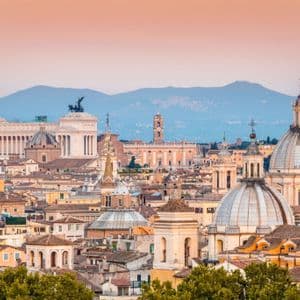 Un paesaggio urbano di edifici storici e cupole al tramonto, con montagne visibili sullo sfondo.