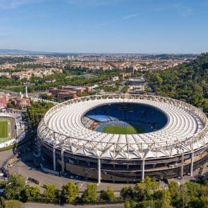 Una vista aerea di un grande stadio sportivo circolare con un tetto bianco, situato accanto a una pista di atletica più piccola ai margini di una città.