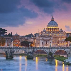 Una basilica a cupola illuminata e un ponte ad arco in pietra che attraversa un ampio fiume al crepuscolo, sotto un cielo nuvoloso e colorato.