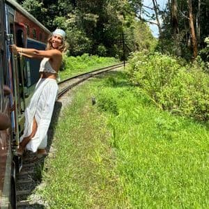 Une femme souriante vêtue de blanc et portant un bandana se penche hors d'un train en mouvement alors qu'il traverse une forêt luxuriante et verte.