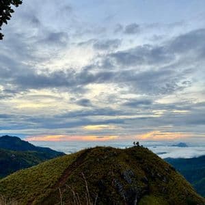 Un pic montagneux verdoyant avec un chemin de terre dominant une vallée remplie de nuages, lors d'un lever de soleil coloré.