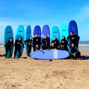 Un gruppo WeRoad in muta posa con le tavole da surf su una spiaggia sabbiosa sotto un cielo azzurro.