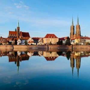 Un paesaggio urbano con edifici storici e campanili che si riflettono perfettamente sull'acqua calma di un fiume sotto un cielo azzurro limpido.