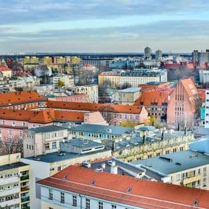 Un panorama cittadino con edifici vari, tetti di tegole rosse e un'imponente cattedrale con due guglie, visibile contro un cielo nuvoloso.