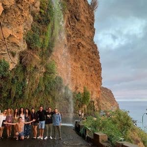 Un gruppo WeRoad in posa su una strada sotto una cascata che scende da una scogliera, con il mare sullo sfondo.
