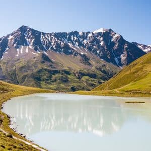 Ein milchig-blauer Alpensee spiegelt eine schneebedeckte Bergkette wider, mit einem gewundenen Pfad entlang seines grasbewachsenen Ufers.