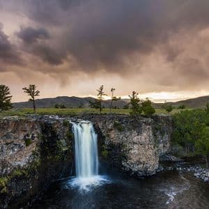 Una cascata si getta da una scogliera rocciosa in un fiume scuro, con verdi pianure e montagne distanti sotto un cielo drammatico e nuvoloso al tramonto.