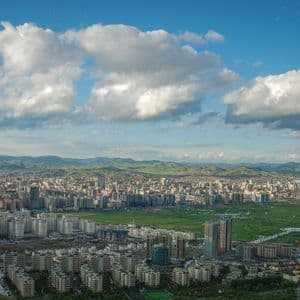 Una vasta città in una valle verde con dolci colline in lontananza, vista dall'alto sotto un cielo parzialmente nuvoloso.