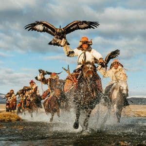 Un gruppo di cacciatori di aquile in tradizionali abiti di pelliccia galoppa a cavallo attraverso un fiume, tenendo le aquile sulle braccia.