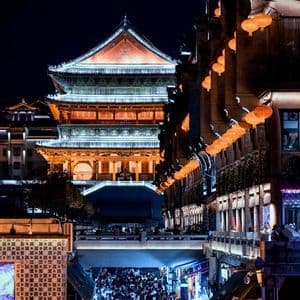 Un grande edificio tradizionale asiatico illuminato si affaccia su una strada cittadina affollata di persone sotto un cielo notturno buio.