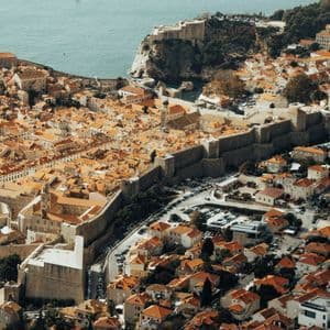 Veduta aerea di una storica città murata con tetti in tegole arancioni, fortificazioni e un piccolo porto sulla costa.