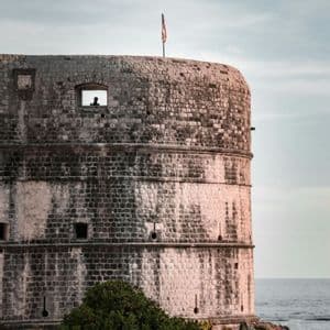 Una torre fortezza in pietra sul mare, con la sagoma di una persona in una finestra e una bandiera sul tetto sotto un cielo nuvoloso.