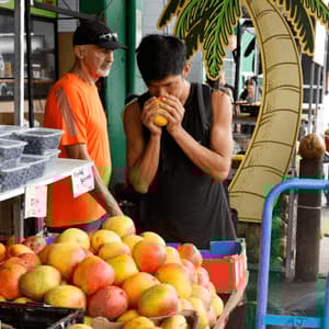 Un uomo in canotta nera annusa un mango giallo a una bancarella di frutta in un mercato all'aperto.