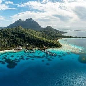 Vista aerea di un'isola tropicale e montuosa con bungalow sull'acqua e una barriera corallina in acque turchesi.
