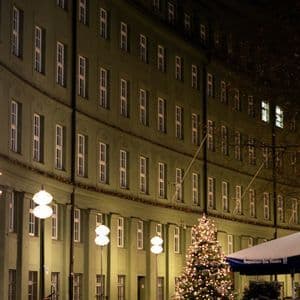 Di notte, un albero di Natale illuminato si erge su una piazza di ciottoli bagnata, davanti a un grande edificio verde e curvo con molte finestre.
