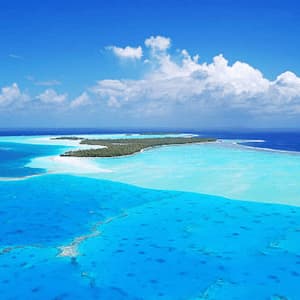 Veduta aerea di un atollo tropicale circondato da una laguna turchese poco profonda e dall'oceano blu intenso, sotto un cielo azzurro con nuvole bianche.
