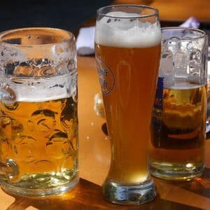 Tres vasos de cerveza diferentes, incluyendo una jarra grande y un vaso alto de cerveza de trigo, están sobre una mesa de madera bajo la luz del sol.