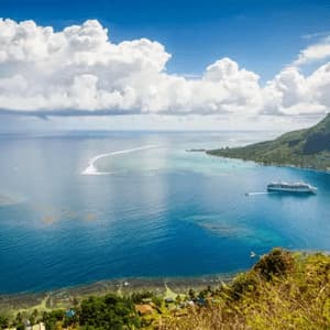 Una vista aerea di una nave da crociera bianca che naviga in una baia turchese accanto a una montagna verde lussureggiante sotto un cielo con grandi nuvole bianche.