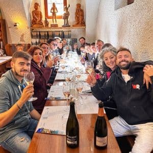 Un viaggio di gruppo WeRoad si gode una degustazione di vini, alzando i calici per un brindisi a un lungo tavolo di legno all'interno di una cantina rustica.