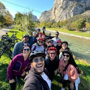 Un gruppo WeRoad in viaggio, con caschi da ciclista, si scatta un selfie su una sponda erbosa del fiume, con ripide montagne sullo sfondo.