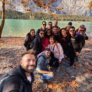 Un gruppo WeRoad sorride per un selfie in una giornata autunnale in riva a un lago con montagne sullo sfondo.