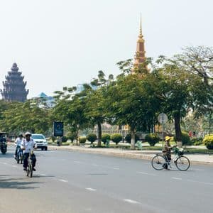 Un'ampia strada cittadina con persone in bicicletta, in scooter e a bordo di tuk-tuk. Alberi verdi costeggiano la strada, con torri ornate sullo sfondo.