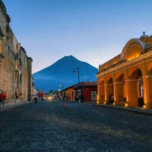 Una calle empedrada flanqueada por edificios coloniales iluminados conduce hacia un gran volcán al atardecer.