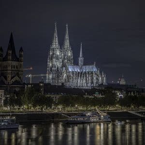 Una grande cattedrale gotica con guglie gemelle è illuminata contro il cielo notturno, vista da oltre un fiume con barche.
