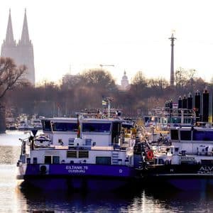 Diverse grandi navi cisterna ormeggiate in un fiume con le guglie gemelle di una cattedrale gotica visibili sullo sfondo nebbioso.