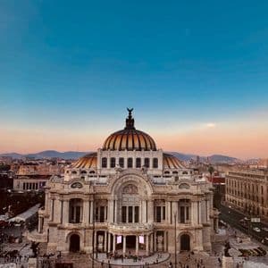 Un palazzo sontuoso e a cupola si erge in una vivace piazza cittadina, sullo sfondo di un cielo al tramonto dai colori vivaci.