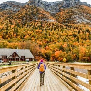 A person with a backpack walks across a wooden bridge towards a large lodge and a mountain covered in colorful autumn foliage.