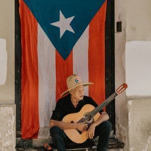 Un anciano con sombrero de paja toca una guitarra sentado en unas escaleras frente a una gran bandera puertorriqueña.